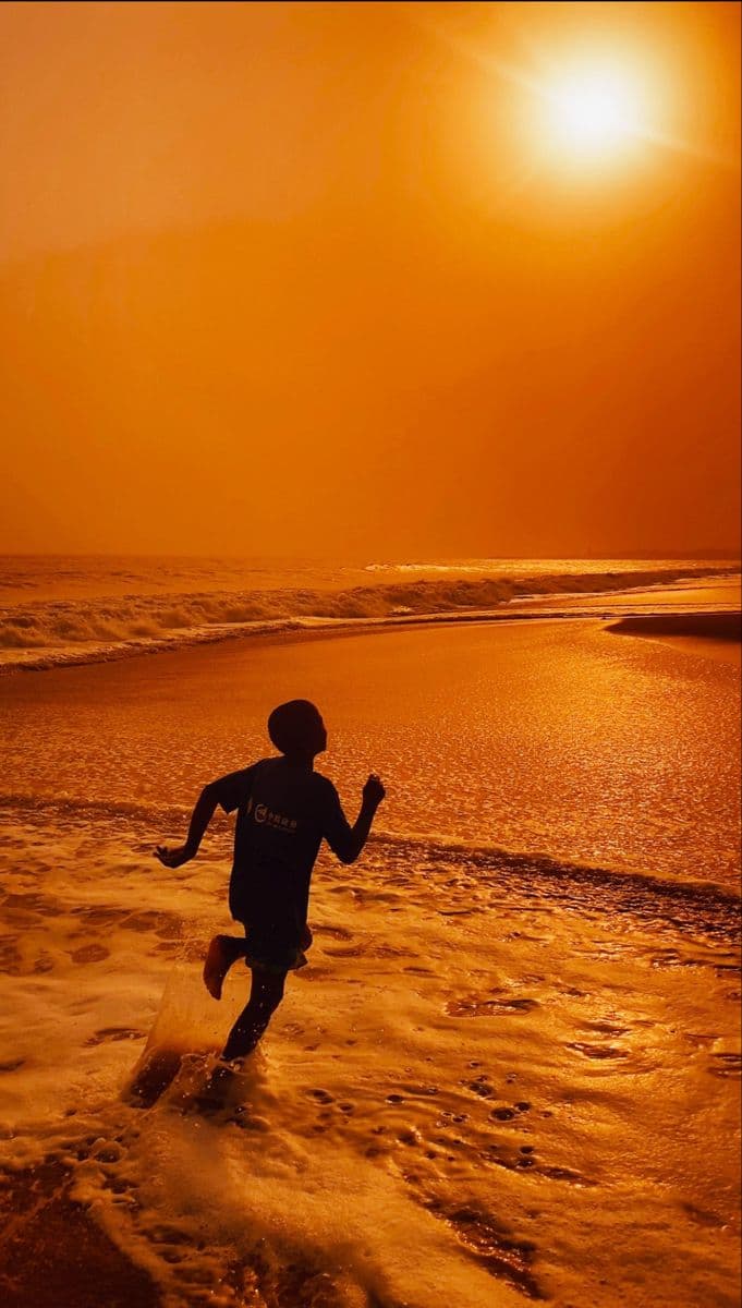 Silhouette of a child running along wet sand under a low orange sun